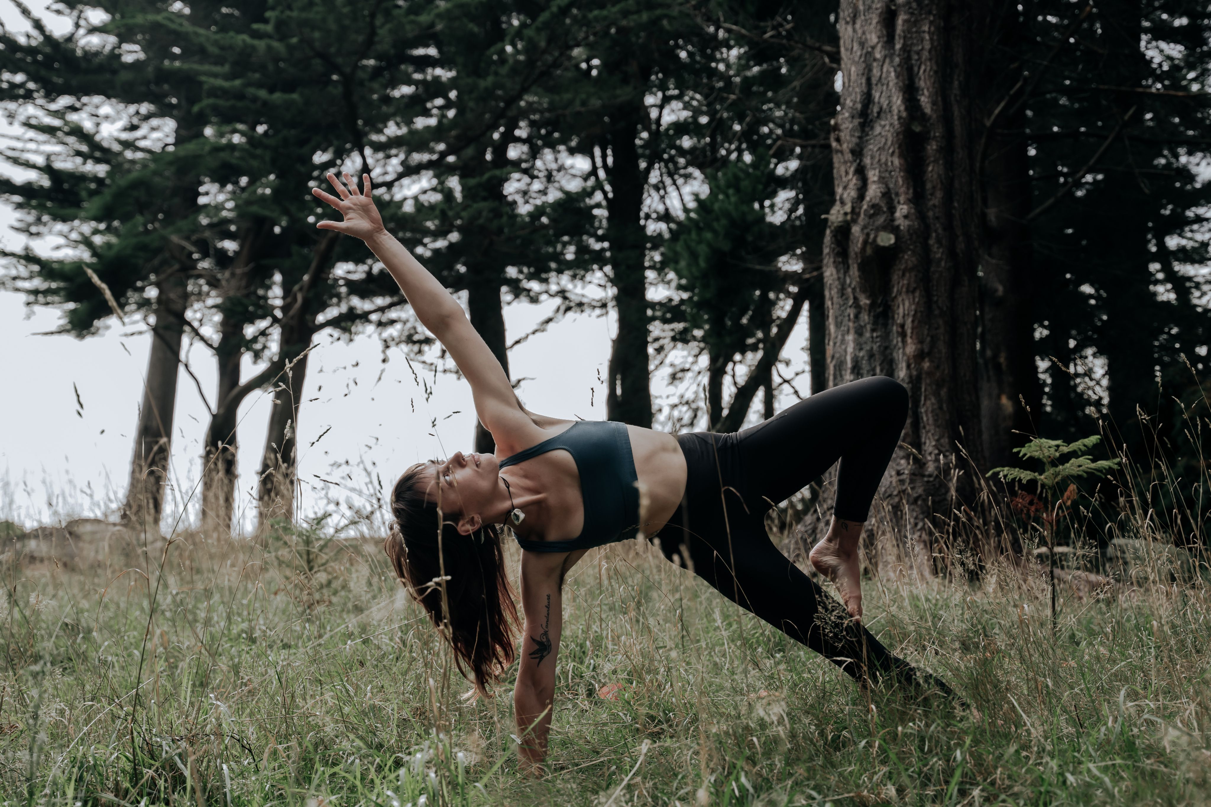 Becca amongst long grass, balancing on her side on one arm and a leg, with the other arm raised towards the sky.